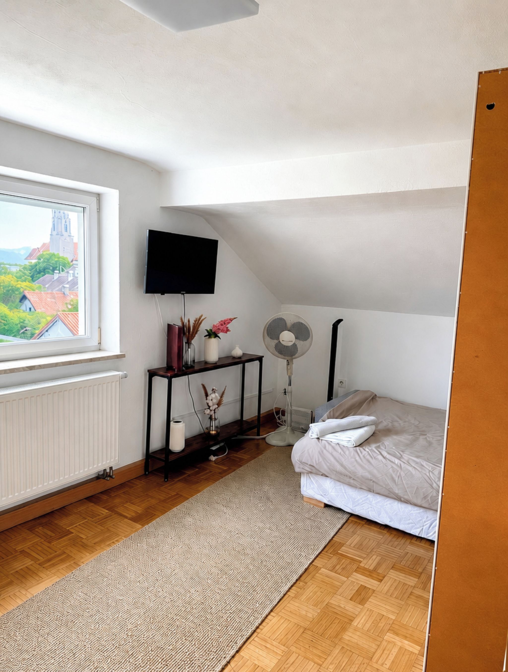 Une chambre à coucher avec un lit, un ventilateur sur pied, un téléviseur fixé au mur, une fenêtre donnant sur des immeubles et un radiateur.