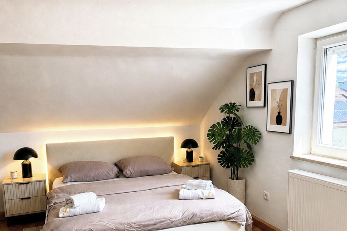 Ett modernt sovrum med en stor säng, sänglampor, en krukväxt och inramade konstverk på väggen.