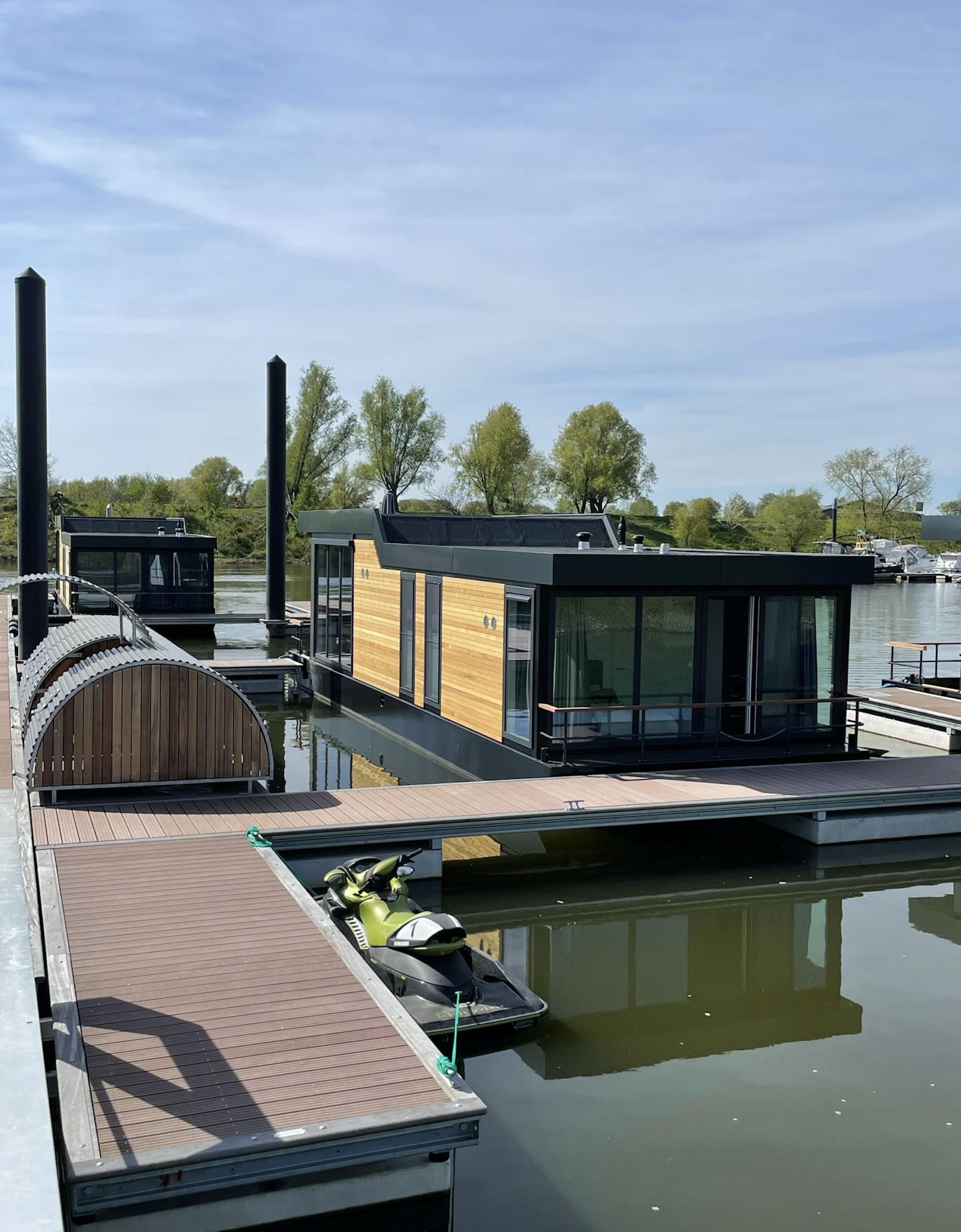 Una casa flotante moderna con terraza de madera, una patineta verde y un cuerpo de agua tranquilo al fondo.