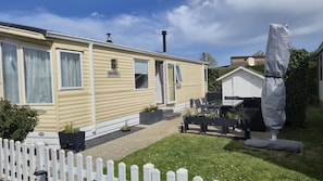 Ein Mobilheim mit einer Terrasse, einem eingezäunten Garten und einem kleinen Geräteschuppen.