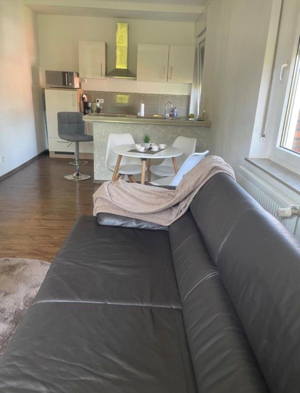 Een moderne woonkamer met een donker leren bank, een klein rond tafeltje met stoelen, en een keuken op de achtergrond.