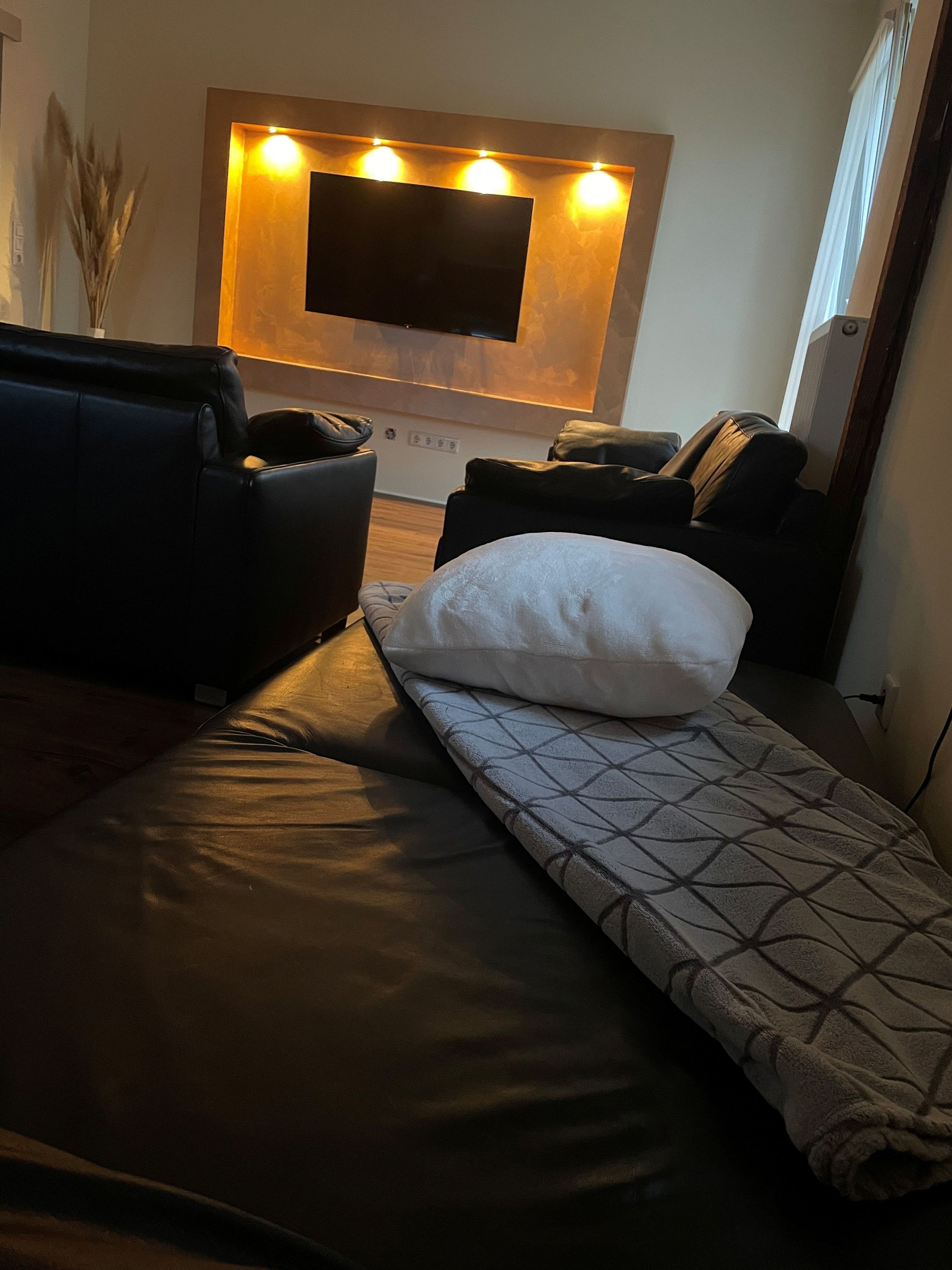 Ein modernes Wohnzimmer mit einer Couch, einem Relaxsessel, einem an der Wand befestigten Fernseher und einer gemusterten Decke auf dem Boden.