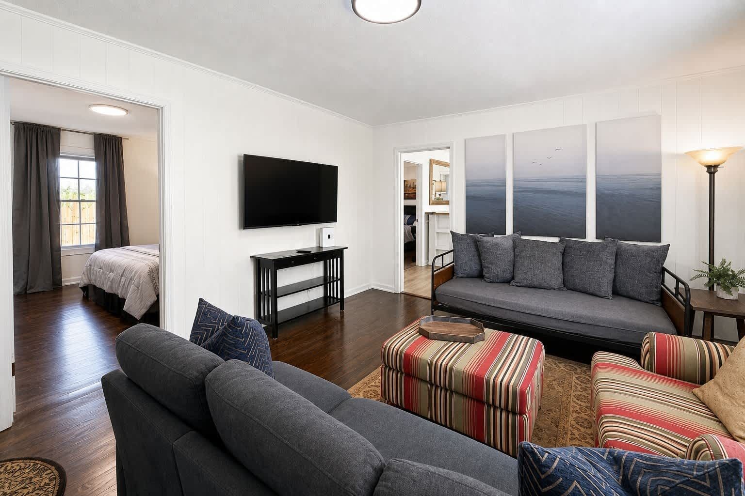 A modern living room with a flat-screen TV, a gray sofa, striped ottomans, and a view of the outdoors.
