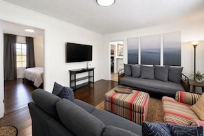 A modern living room with a flat-screen TV, a gray sofa, striped ottomans, and a view of the outdoors. - King Bed Cottage, close to Swamp Rabbit + Downtown (Greenville)