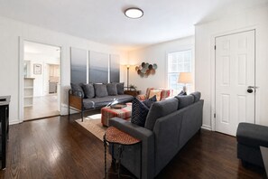 A modern living room with a grey sofa, a coffee table, and a side table. There is a door on the right and a window with blinds on the left. - King Bed Cottage, close to Swamp Rabbit + Downtown (Greenville)