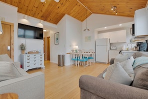 Ein modernes Wohnzimmer mit Holzboden, ein Essbereich mit Tisch und Stühlen sowie eine Küche mit weißen Schränken und Geräten.