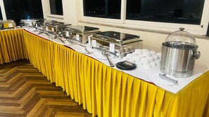 A buffet setup with multiple serving stations, a large pot on the right, and a window in the background. - Hotel Ambaari Inn (Mandya)