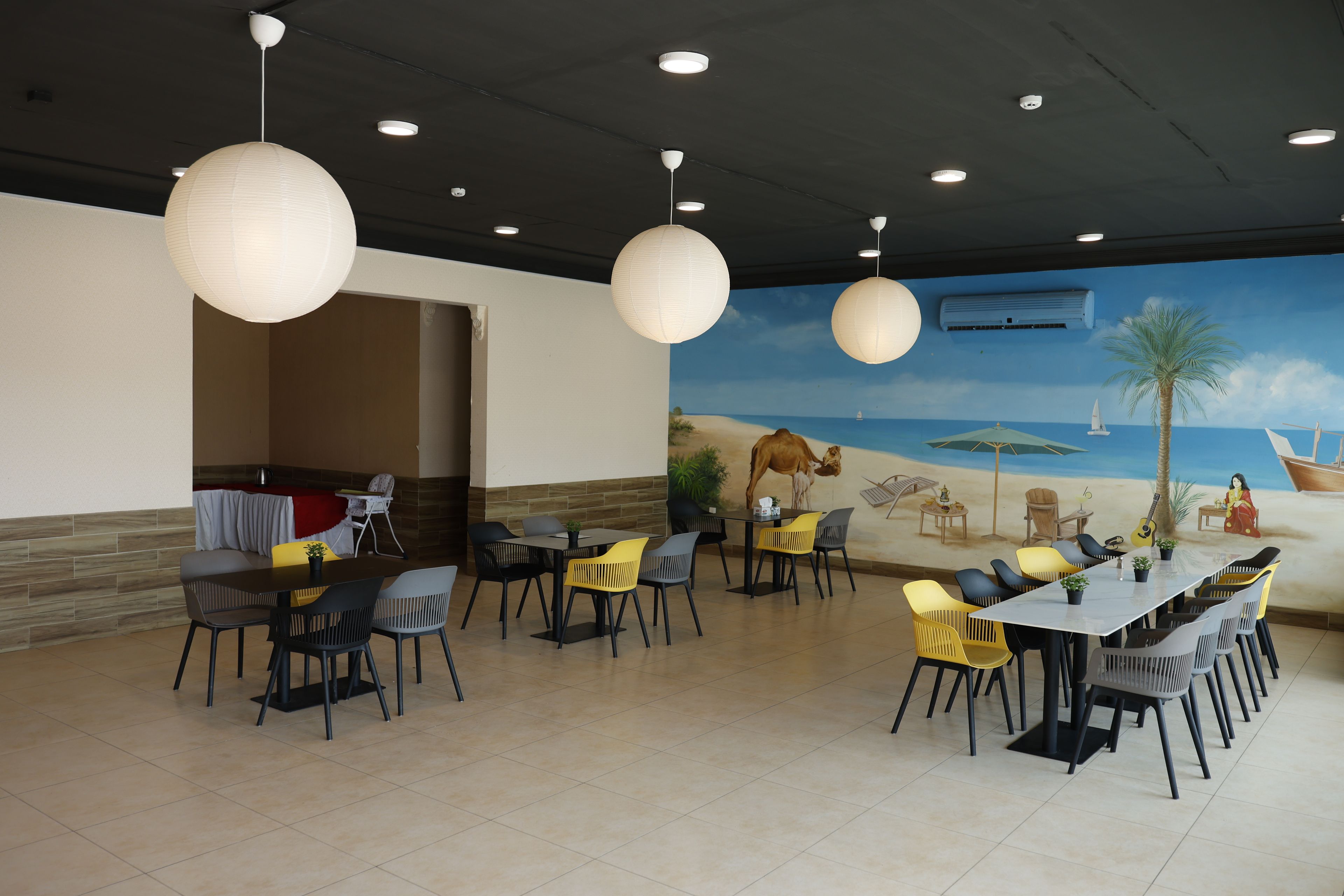 A restaurant with multiple tables and chairs, a mural of a beach scene, and ceiling lights.