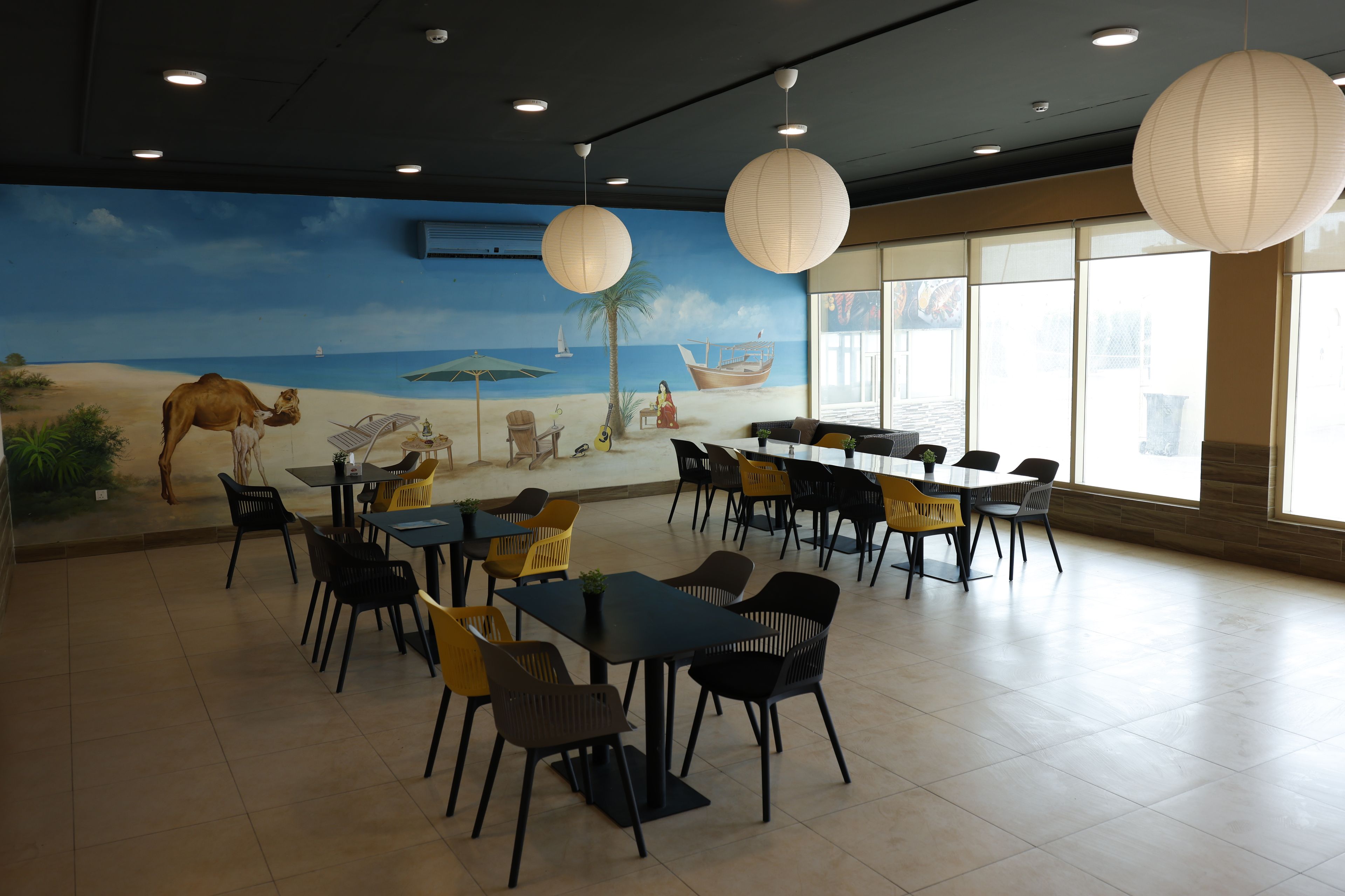 A dining area with tables and chairs, a mural of a beach scene, and large paper lanterns.