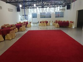 Un salón amplio con alfombra roja, mesas dispuestas en filas con manteles rojos y sillas con cintas doradas.