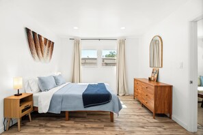 Une chambre à coucher comprenant un lit, une table de chevet, une commode et une grande fenêtre avec des rideaux.