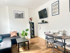 Een moderne woonkamer met een bank, salontafel, eettafel met stoelen, een aan de muur bevestigde televisie en een boekenkast met decoratieve objecten.
