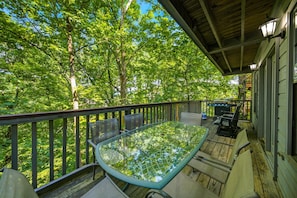 Eine überdachte Terrasse im Freien mit einem Glastisch und Stühlen, umgeben von einem Holgitter, mit Blick auf eine bewaldete Landschaft.