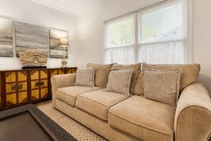 A beige sofa with multiple cushions, a wooden cabinet with drawers, and a large window with blinds. - Cozy Greenville Cottage | 2 Queens + Near Downtown (Greenville)