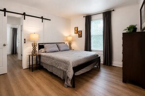 Ein Schlafzimmer mit einem Bett, Nachttischlampen, einem Fenster mit Vorhängen und einer Tür zu einem weiteren Raum.