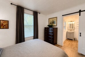 A bedroom with a bed, a dresser, a window with curtains, a painting, and a door leading to a bathroom. - Cozy Greenville Cottage | 2 Queens + Near Downtown (Greenville)