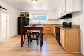 A kitchen with wooden cabinets, a black refrigerator, a wooden dining table with stools, and a window with a view of a blue house. - Cozy Greenville Cottage | 2 Queens + Near Downtown (Greenville)