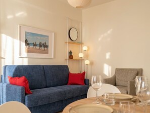 A living room with a blue sofa, red cushions, a dining table with plates and glasses, a wall-mounted clock, and a framed picture of horseback riders. - Versilia escape | 80 meters to the beach (Camaiore)