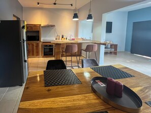 A modern kitchen with a refrigerator, oven, and wooden cabinets. There is a dining area with a wooden table, two chairs, and a small rug. - Newly renovated one-bedroom, one-bath ground unit at Turquoise Condominiums (Playas Del Coco)