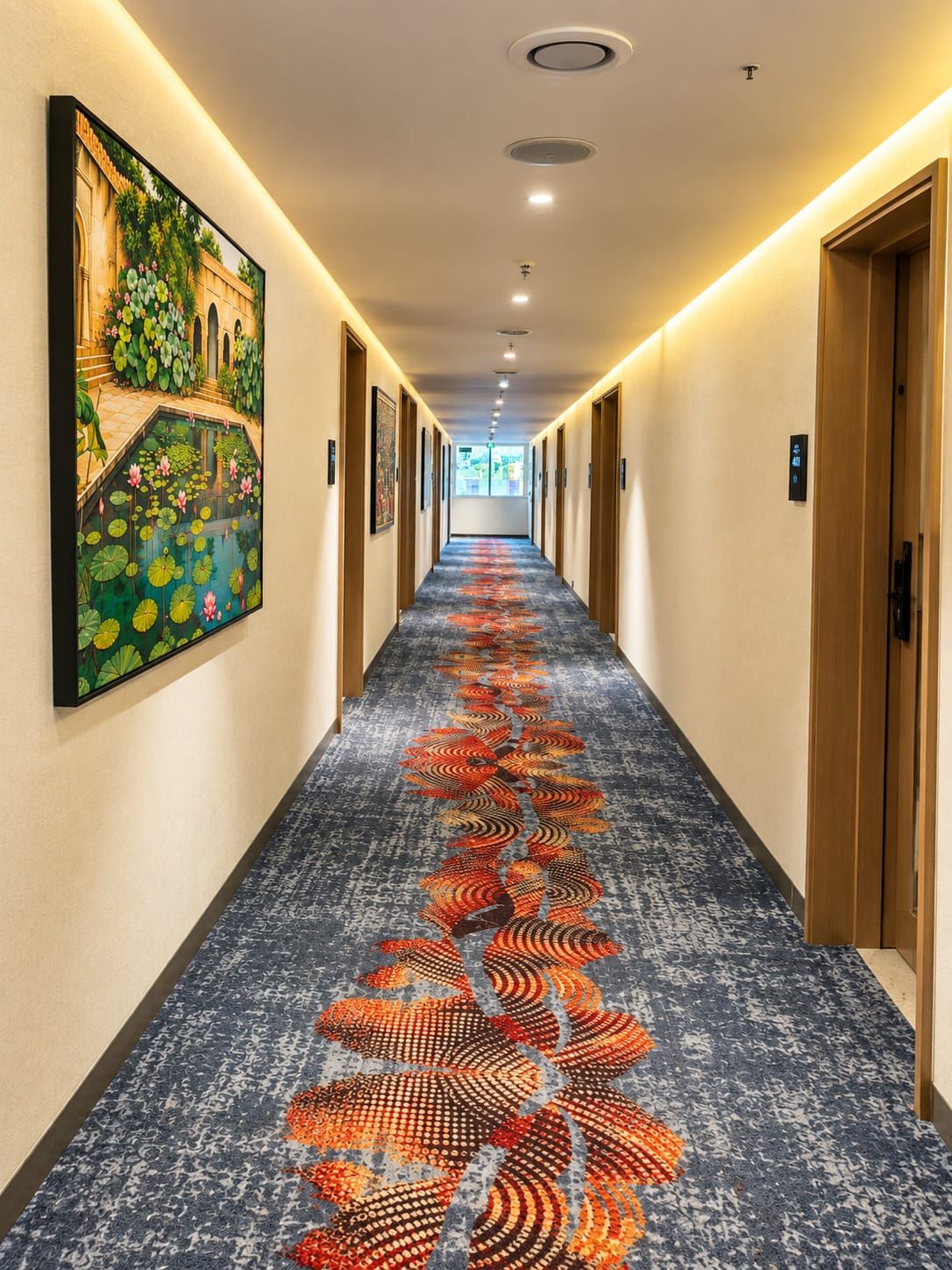 A hotel corridor with a patterned carpet, doors on both sides, and a painting on the left wall.