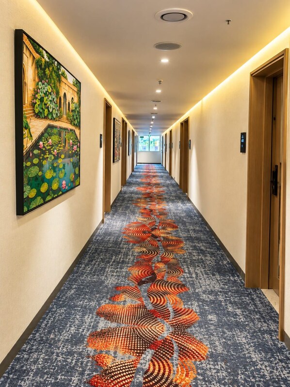 A hotel corridor with a patterned carpet, doors on both sides, and a painting on the left wall.