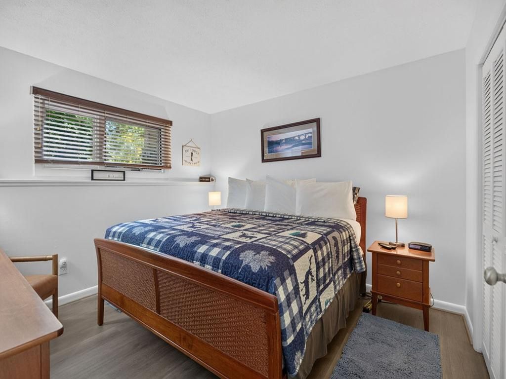 Une chambre à coucher avec un lit en bois, une table de chevet, une commode, une fenêtre avec des stores, un tableau encadré au mur et une porte avec une poignée.