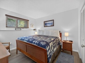 Een slaapkamer met een houten bed, een nachtkastje, een raam met zonwering, een ingelijste foto aan de muur en een deur met een handgreep.