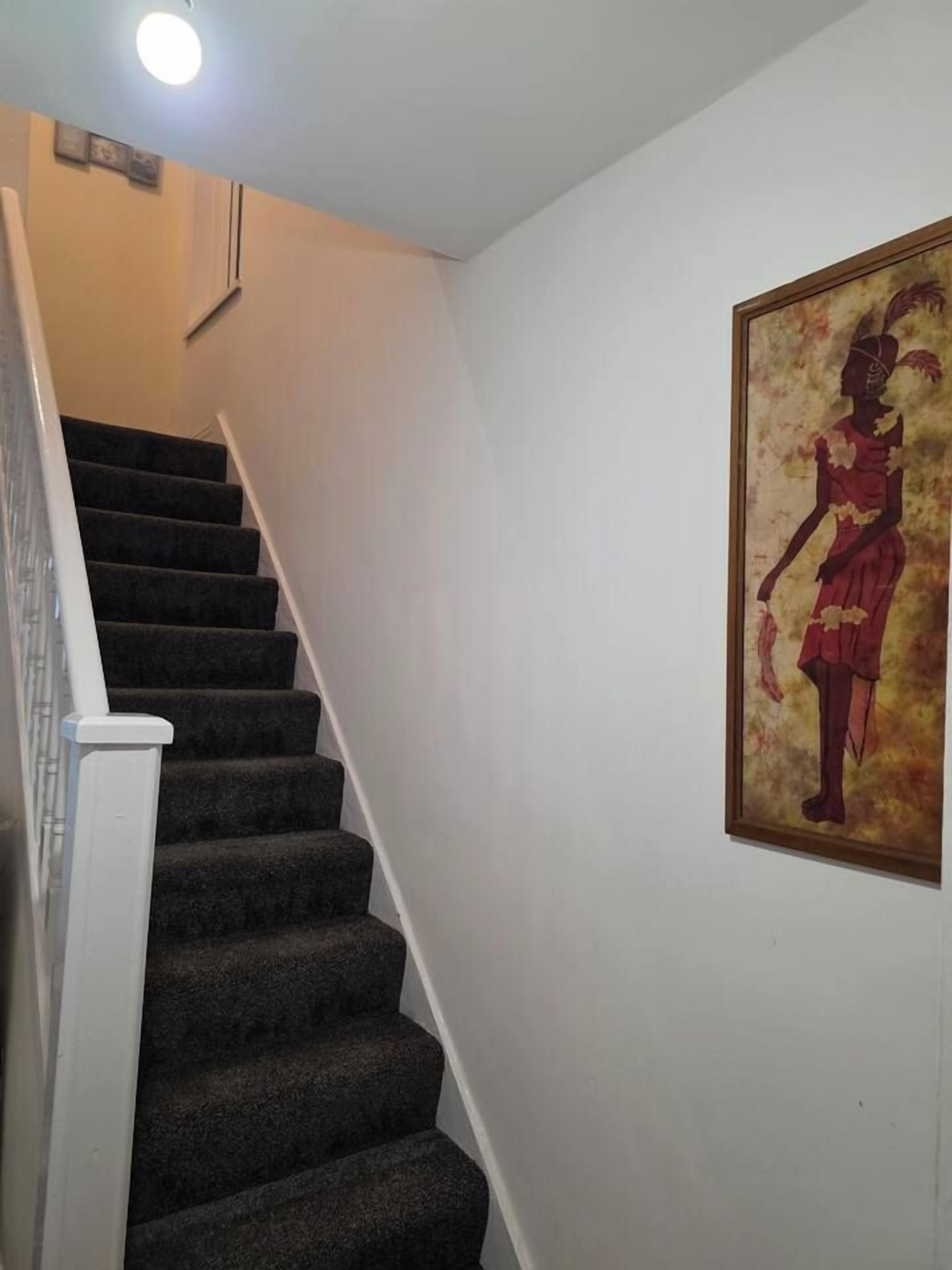 Un escalier dont les marches sont recouvertes de moquette et qui possède une rampe blanche, menant à un palier orné d’une œuvre d’art encadrée au mur.