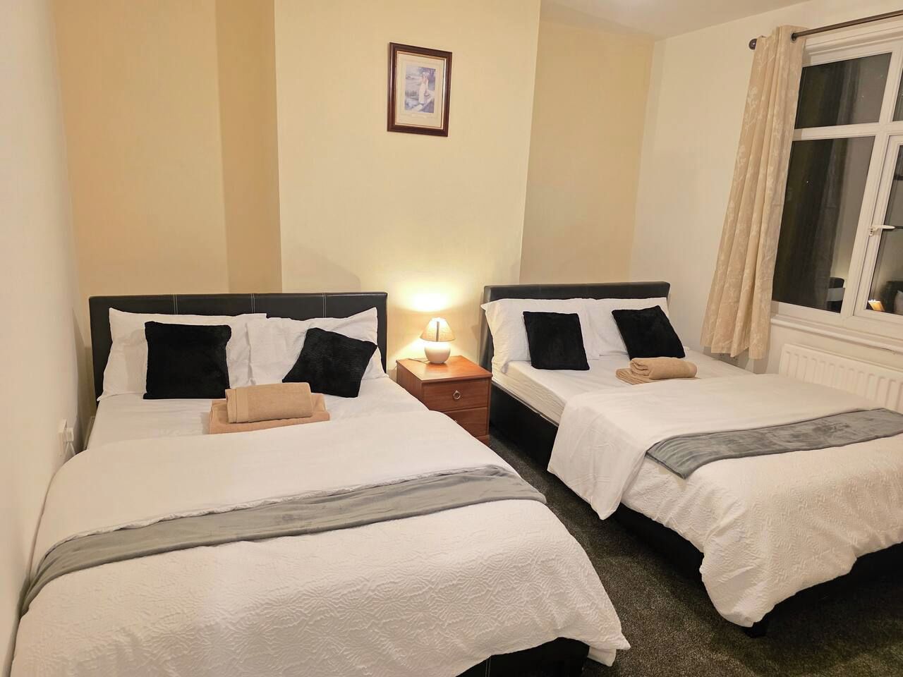 A hotel room with two beds, a nightstand with a lamp, and a window with curtains.