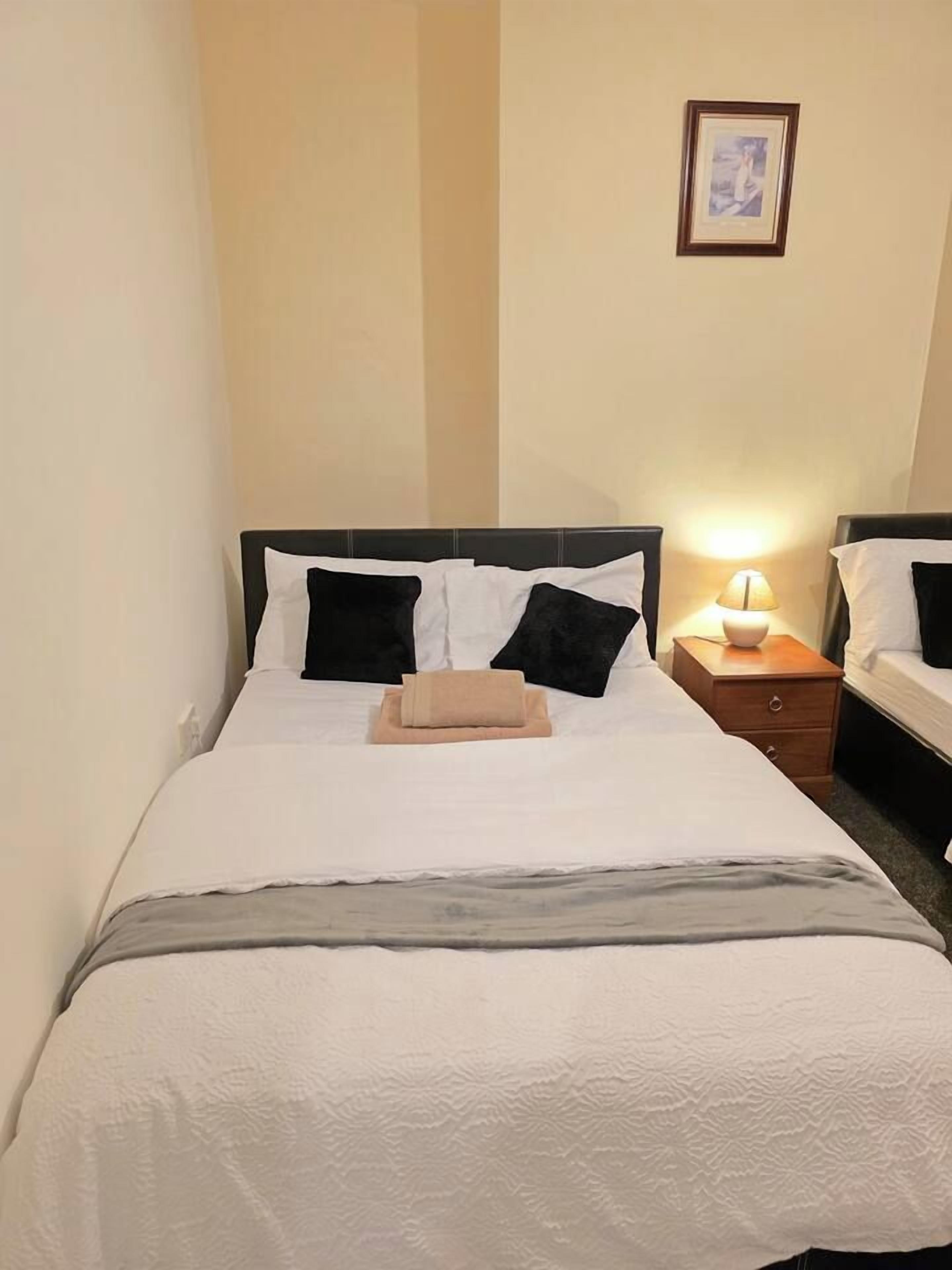 A single bed with a white and gray bedspread, black and white pillows, and a beige towel. There is a nightstand with a lamp and a framed picture on the wall above the bed.