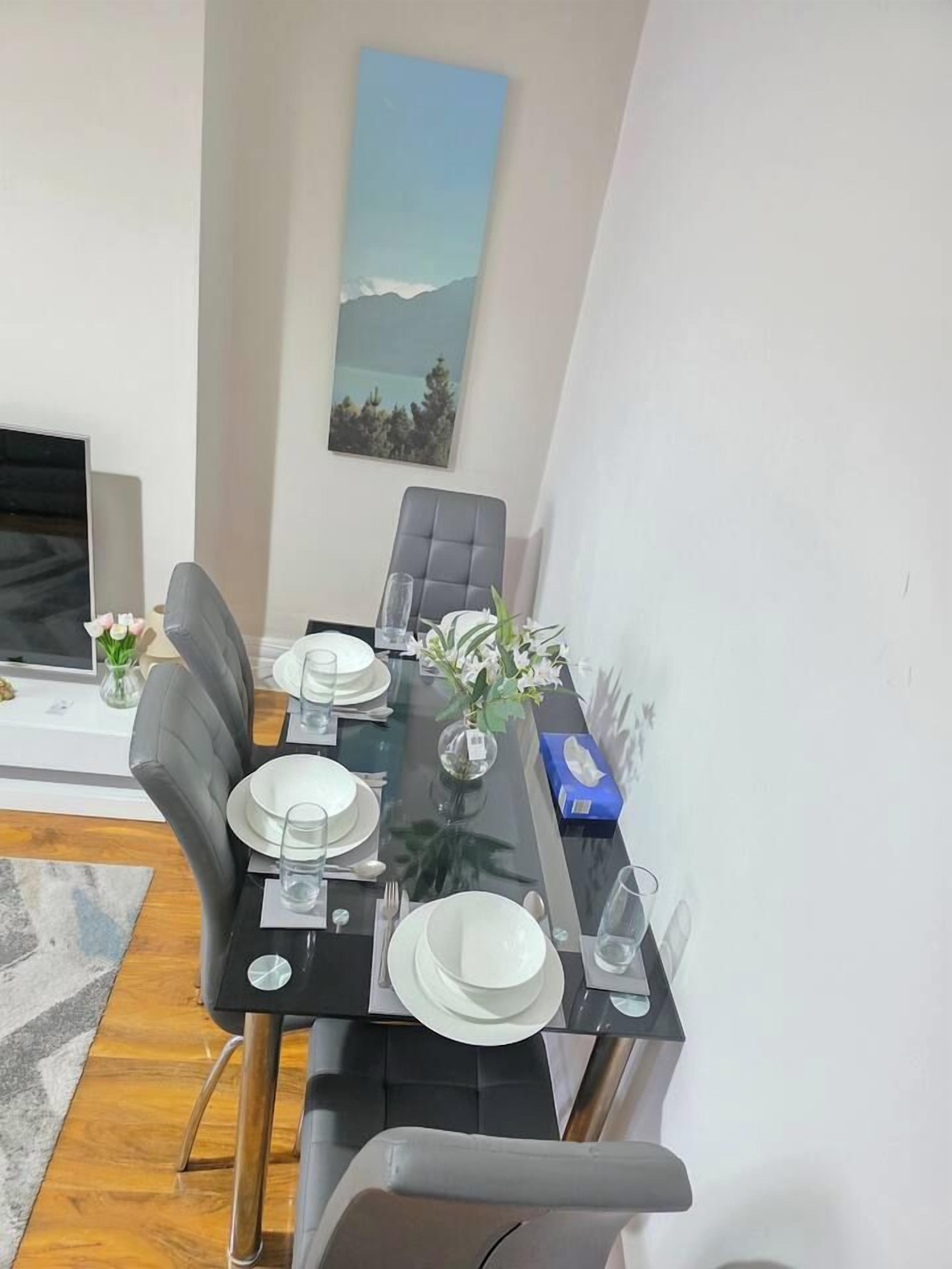 A dining area with a glass table set for four, grey upholstered chairs, and a painting on the wall.