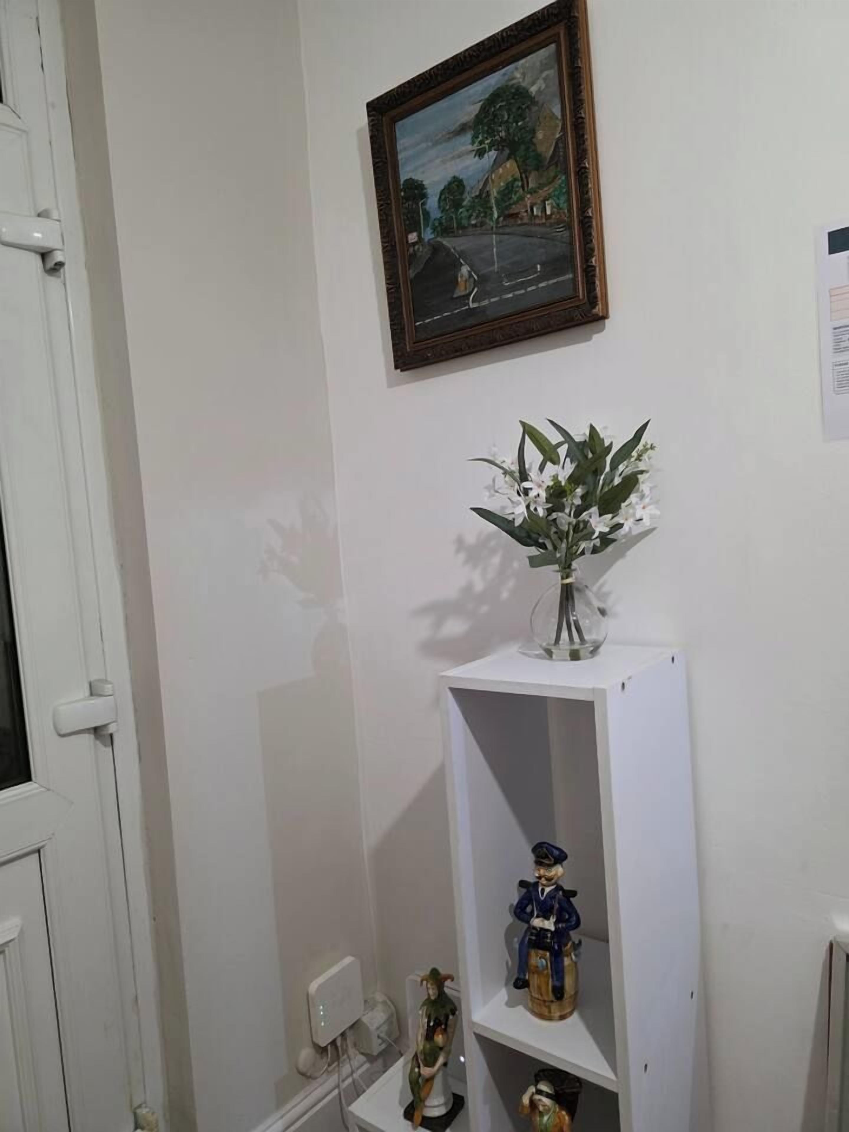 Une pièce avec une porte blanche, une étagère blanche ornée d’un vase de fleurs, un tableau encadré et une petite statue.