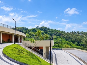 A modern building with a curved staircase, a flat roof, and a view of a lush green hill.