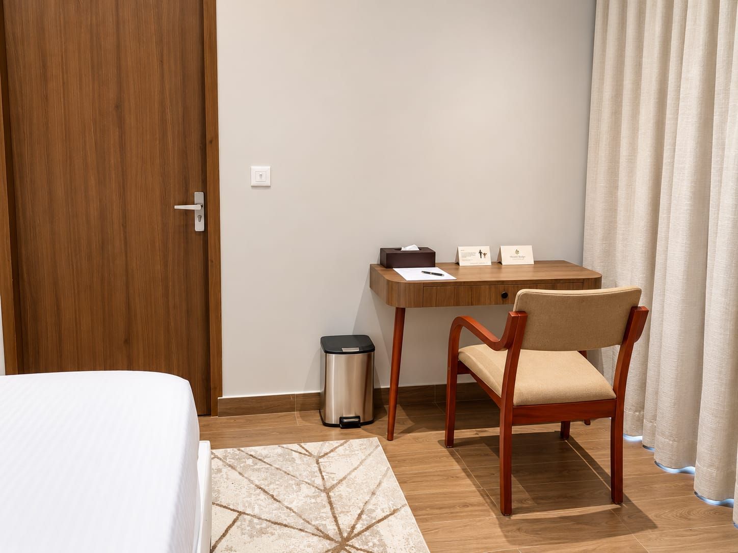 Ein Hotelzimmer mit Holztür, Schreibtisch mit Stuhl, Mülleimer und Bett mit gemustertem Teppich.