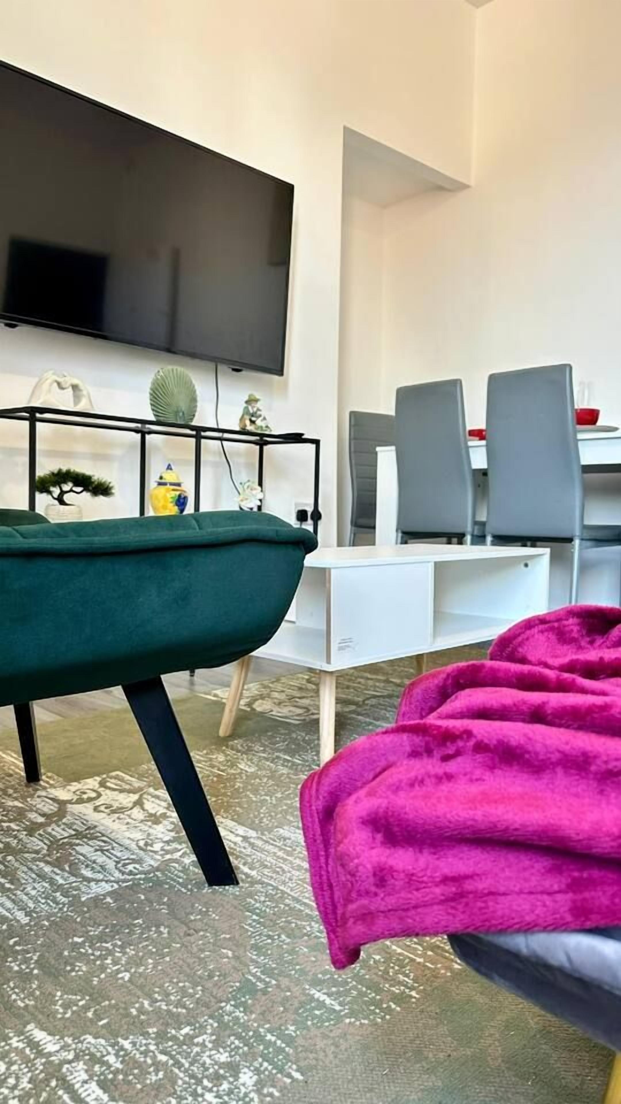 A modern living room with a flat-screen TV mounted on the wall, a green sofa, and a dining area with grey chairs and a white table.