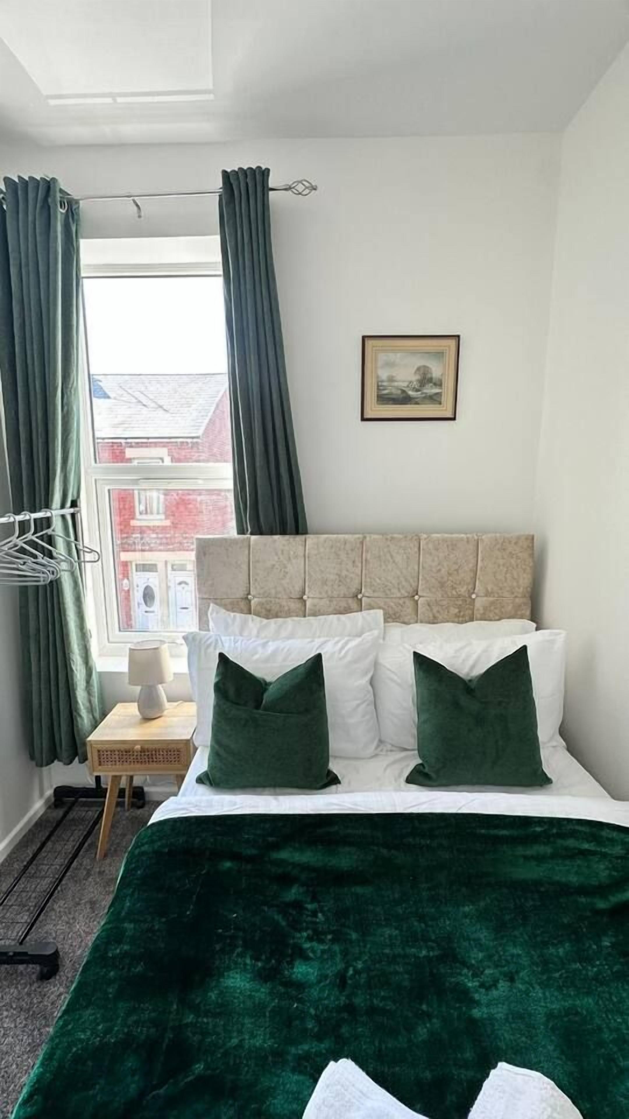Une chambre à coucher avec un lit, une fenêtre avec des rideaux, une table de chevet avec une lampe et un tableau encadré au mur.