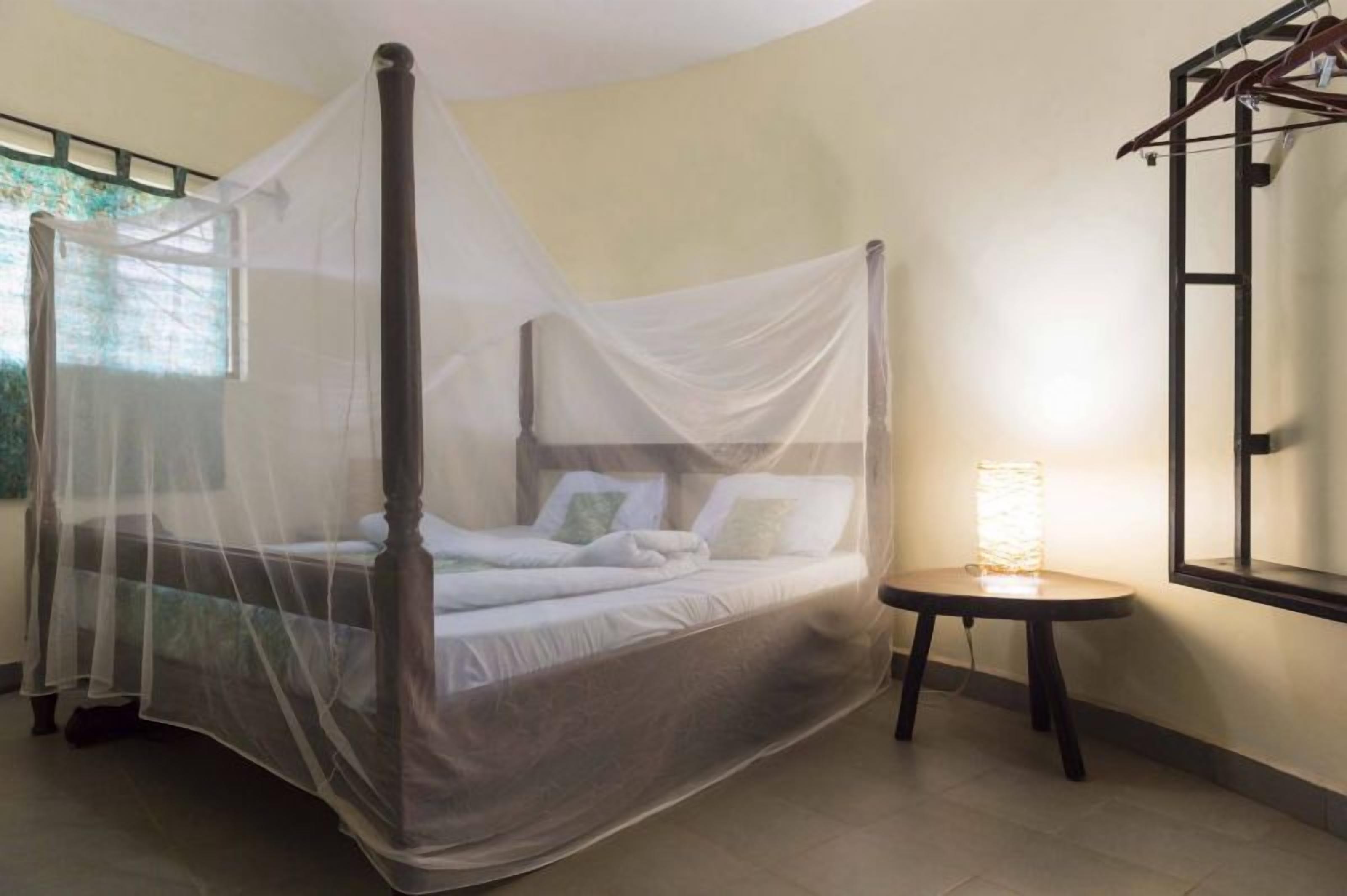 Una cama con dosel y cuatro columnas, una mesita de noche con lámpara, y una ventana con vistas a zonas verdes.