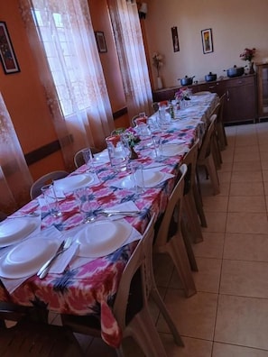 Uma sala de jantar com uma mesa comprida posta para refeição, cadeiras ao redor e utensílios de cozinha sobre a mesa.