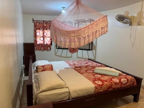 Un dormitorio con cama a dosel, una mesita de noche, un ventilador de pared y una ventana con cortinas.