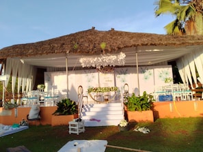Espaço para eventos ao ar livre com telhado de palha, cortinas brancas e decoração floral.