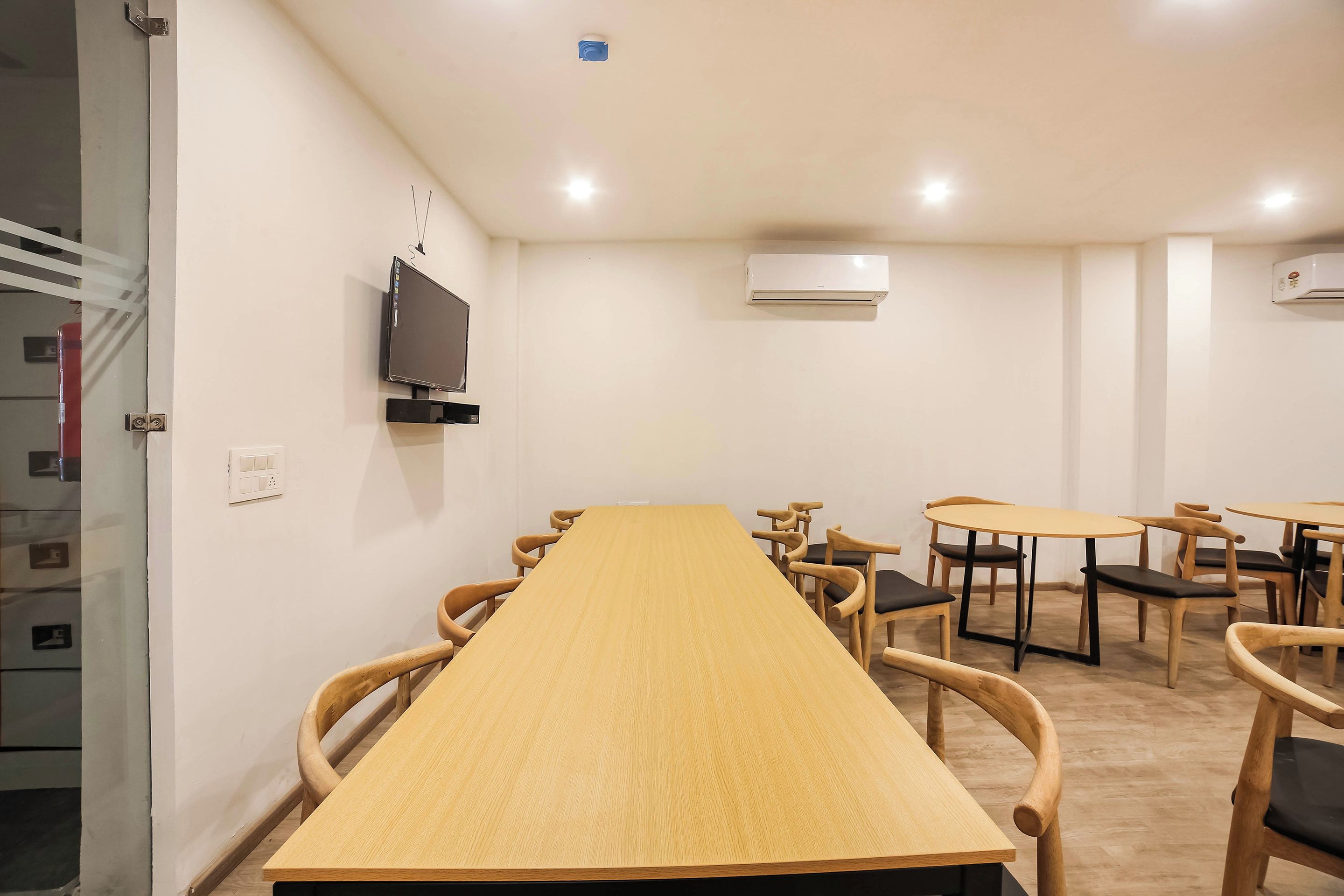 A conference room with a long table, multiple chairs, a flat-screen TV mounted on the wall, and two air conditioning units.