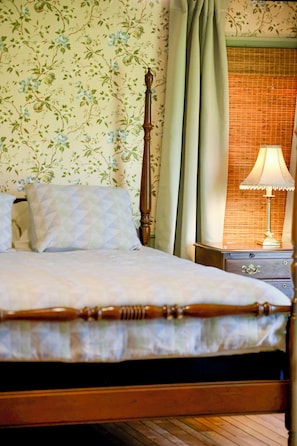 Una cama con estructura de madera, una mesita de noche con lámpara, y una ventana con vista a un muro de ladrillo.