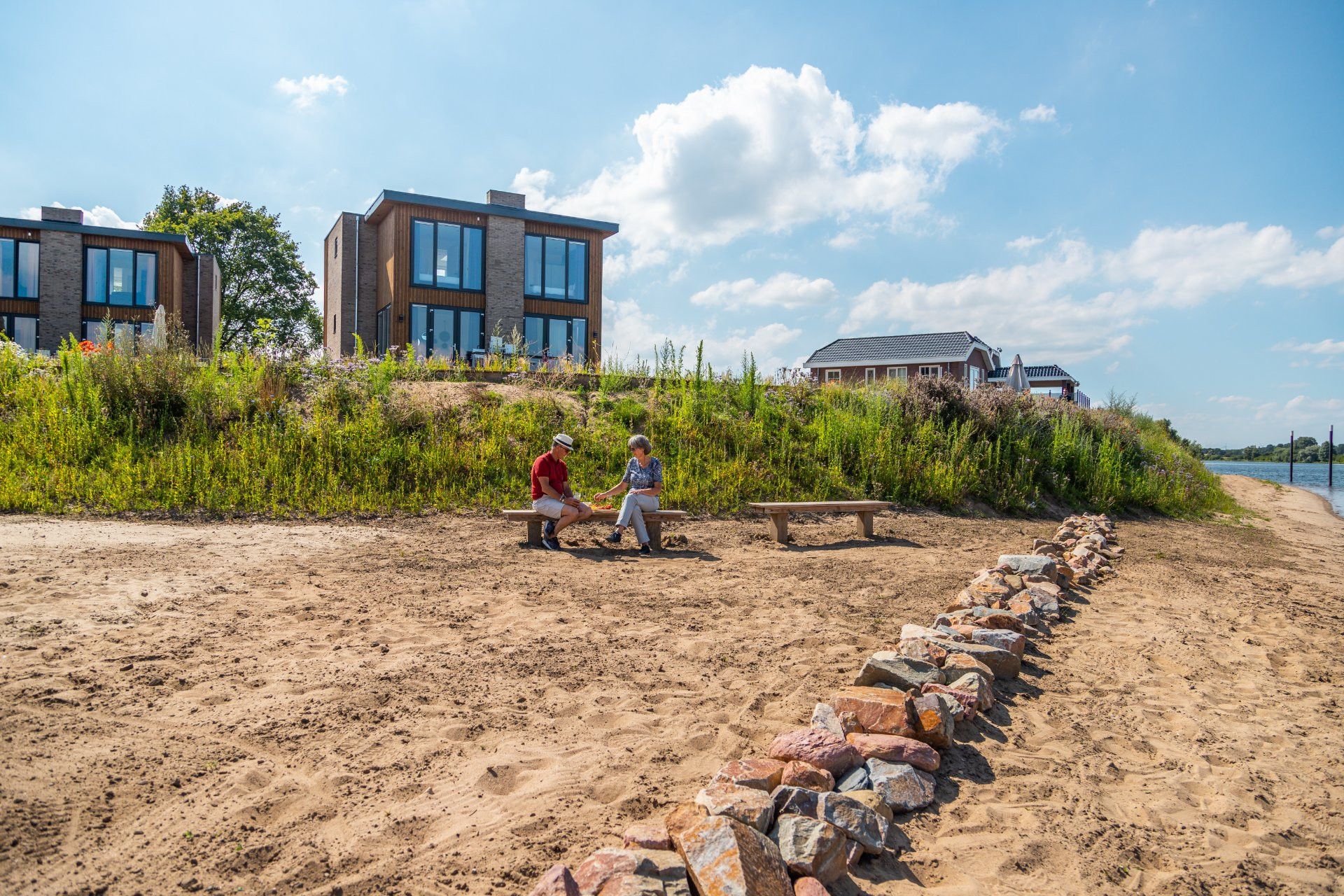 Une plage de sable avec un muret en pierre, deux bancs et deux personnes assises. Au fond, on aperçoit des immeubles modernes dotés de grandes fenêtres.