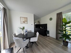 A modern dining area with a round table, two grey chairs, a potted plant, and a kitchen in the background. - Vacation Park Aan de Maas - Weide Lande with a hot tub (Kerkdriel)