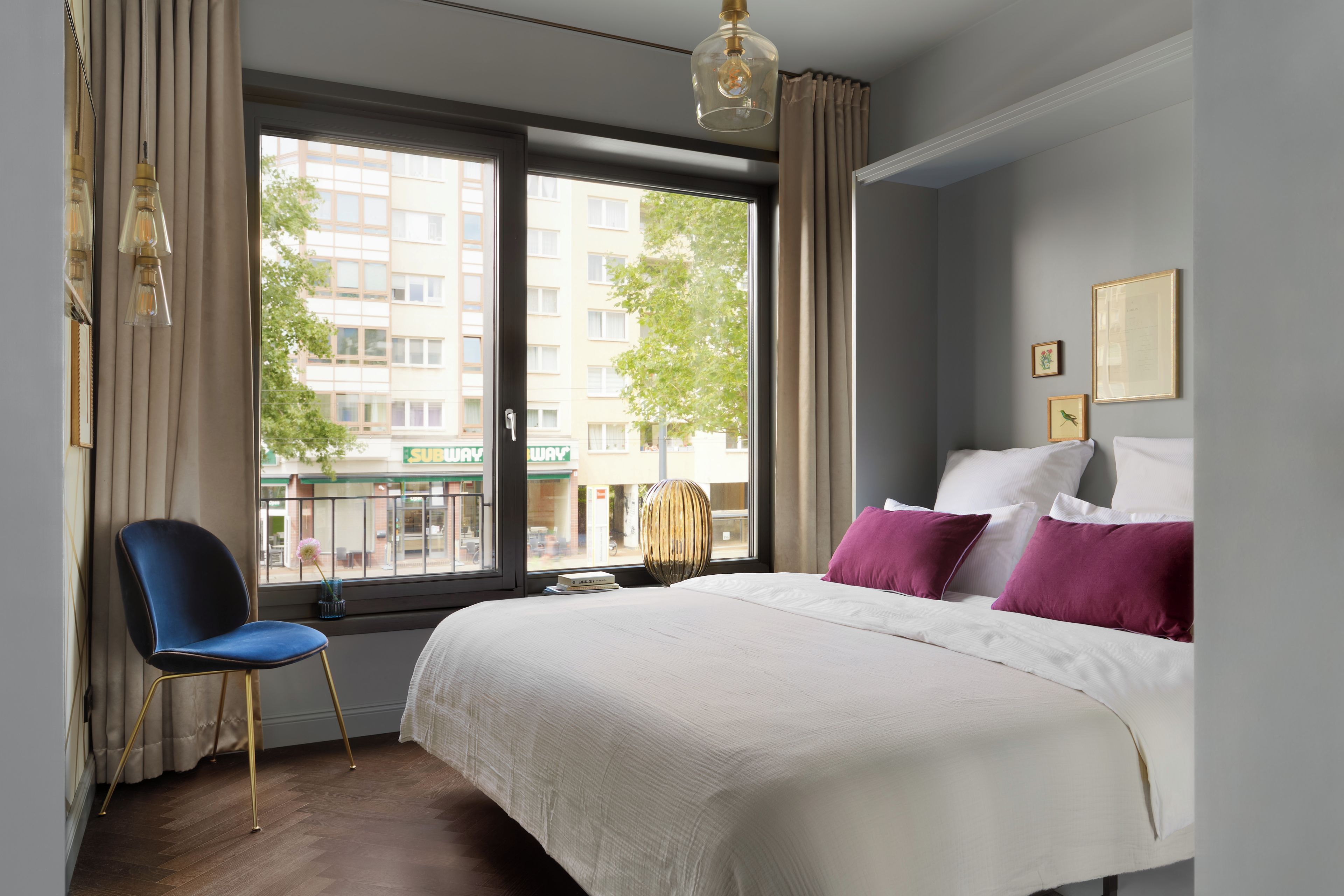 Ein Hotelzimmer mit Bett, Sessel, Fenster mit Blick auf Gebäude und einer Wand mit gerahmten Bildern.