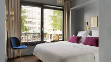 Ein Hotelzimmer mit Bett, Sessel, Fenster mit Blick auf Gebäude und einer Wand mit gerahmten Bildern.