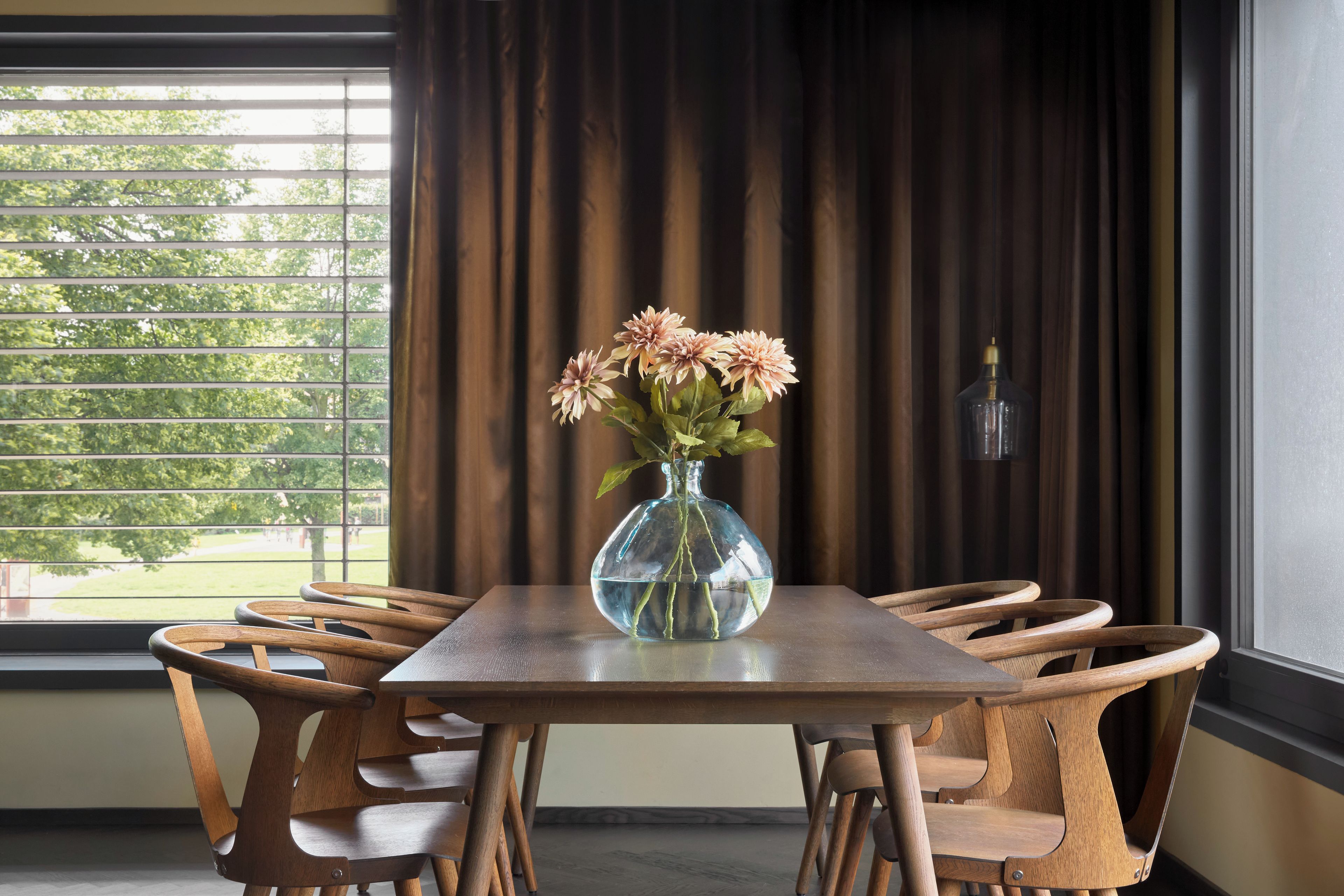 Ein Esstisch mit sechs Holzsesseln, einer Blumenvase und Blick auf Grün durch ein Fenster mit Jalousien.