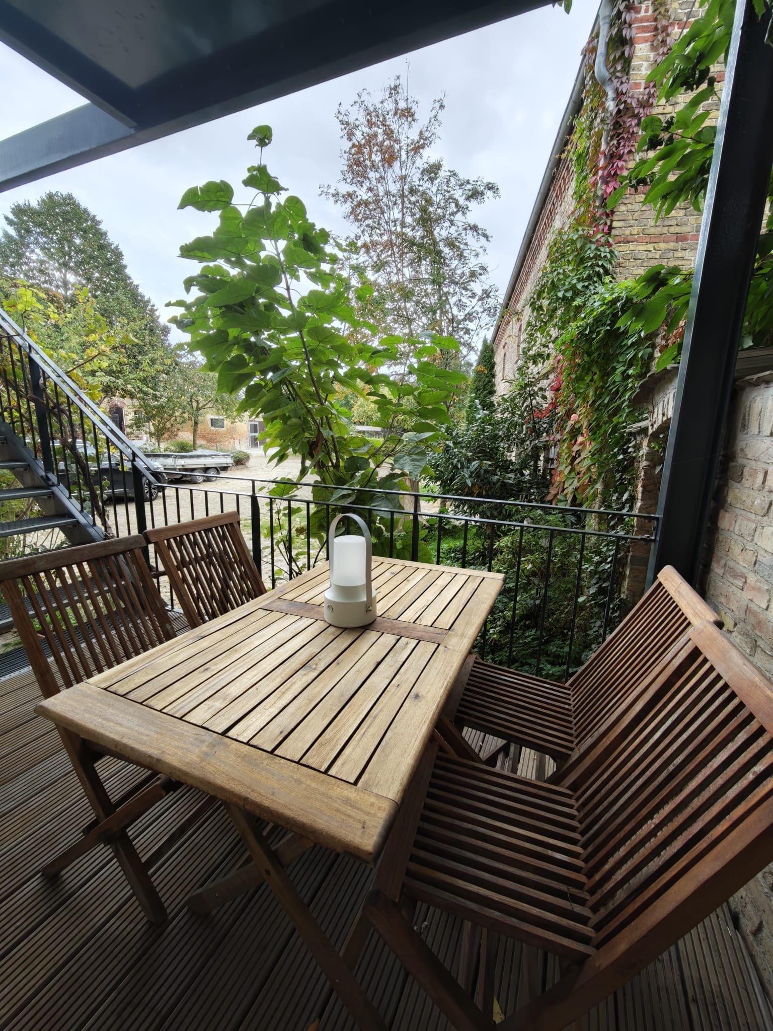 Mesa y sillas de madera en una terraza al aire libre, con vista a un edificio de ladrillo y follaje.