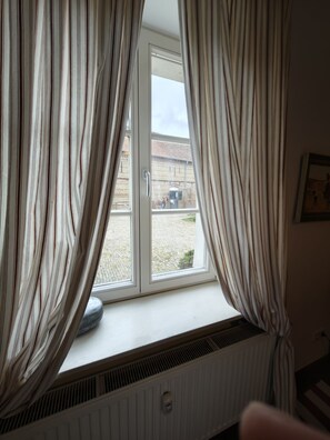 Finestra amb cortines, un radiador a sota i vista d'un edifici i una tanca a l'exterior.