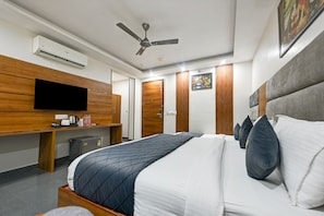 Habitación de hotel con una cama, televisión, ventilador de techo y un pequeño escritorio con tetera y tazas.